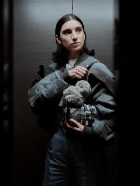 Woman in an elevator holding a gray plush companion on a silver bag