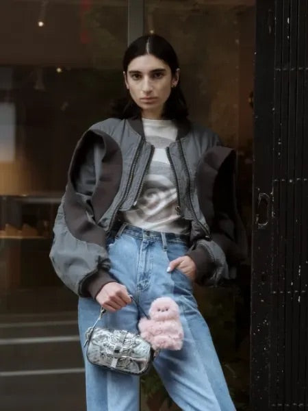 Woman in jeans and a bomber jacket with a pink plush companion on a small silver bag