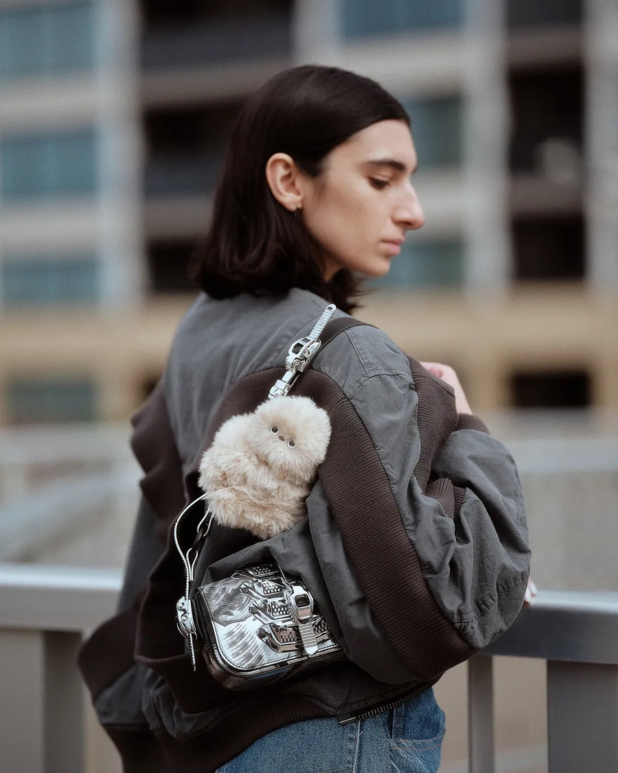 Woman in profile on a rooftop with a cream plush companion on a silver bag strap